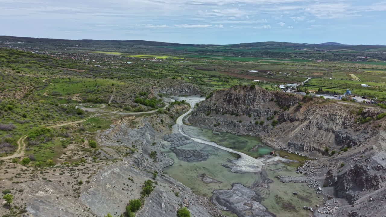 Landskapsbilde av et steinbrudd med en elv og omkringliggende grønne jorder i bakgrunnen på en klar dag.