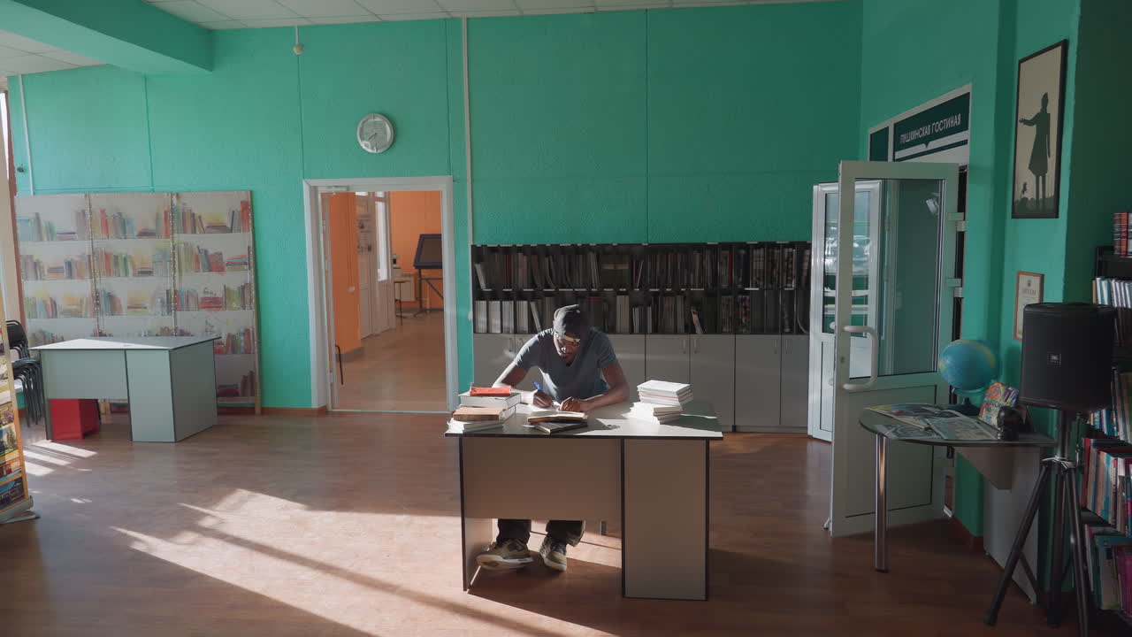 Wide interior view of student in cap studying at desk under sunlight, writing notes between stacks of books in bright quiet library with turquoise walls, open doors, wooden floor, shelves, clock