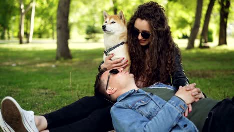 cheerful young man is lying on the grass in the park with his head on his wife's legs while attractive smiling woman is talking to him and caressing well-bred dog.