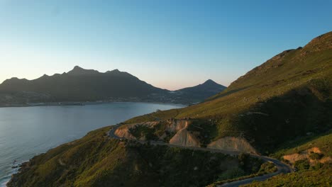 Cars-driving-on-Chapmans-Peak-Road-at-sunset-with-Hout-Bay-in-background,-aerial