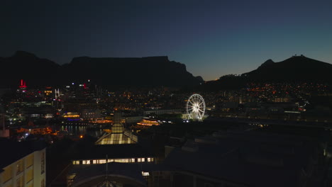 Rückwärts-Fliegen-über-Beleuchtetes-Großes-Gebäude-Des-Einkaufszentrums.-Riesenrad,-Das-In-Die-Nacht-Leuchtet.-Kapstadt,-Süd-Afrika