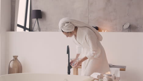 woman in bathrope with a towel in her head opening the tap for fill the bathtub