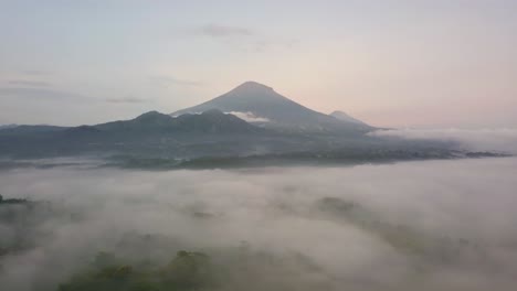 Mystic-Aerial-forward-flight-over-tropical-landscape-covered-with-fog-during-sunny-and-cloudy-day---Massive-Mount-Sumbing-in-background