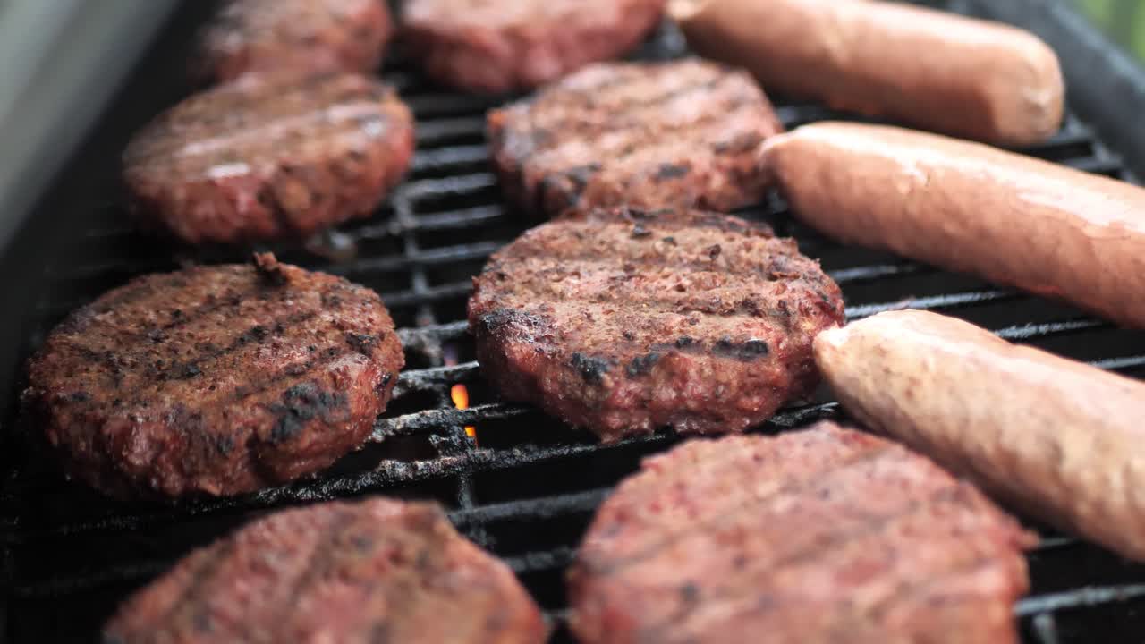 Riprese ravvicinate con panoramica su hamburger e salsicce di carne alternativa sulla griglia con fiamme mentre il coperchio si chiude, girato in slow motion 4k.