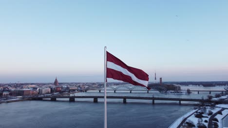 Latvian-red-and-white-flag-fluttering-in-wind-on-pole-with-Riga-city-in-background