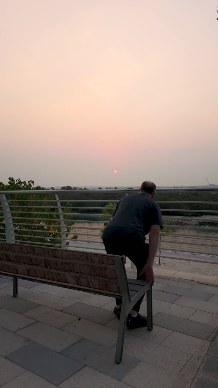 A senior man sits on a park bench at sunrise in slow motion. Captured in vertical video, this serene moment offers a peaceful start to the day, ideal for mobile viewing