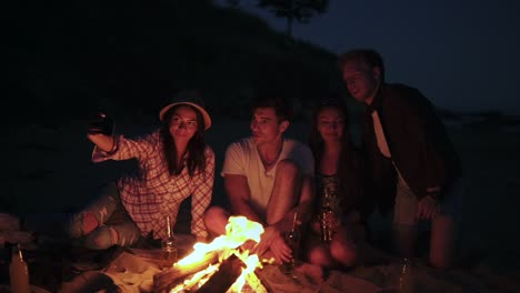 picnic de jóvenes con hoguera en la playa por la noche. amigos alegres tomando fotos en el teléfono. disparo en cámara lenta