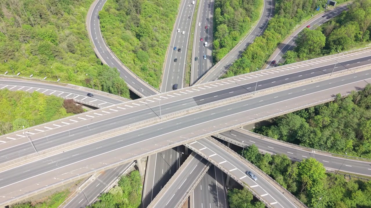 An aerial view shows a busy multi-level highway interchange surrounded by green trees. Cars move along the numerous concrete roads and ramps during daytime, showcasing complex traffic infrastructure.