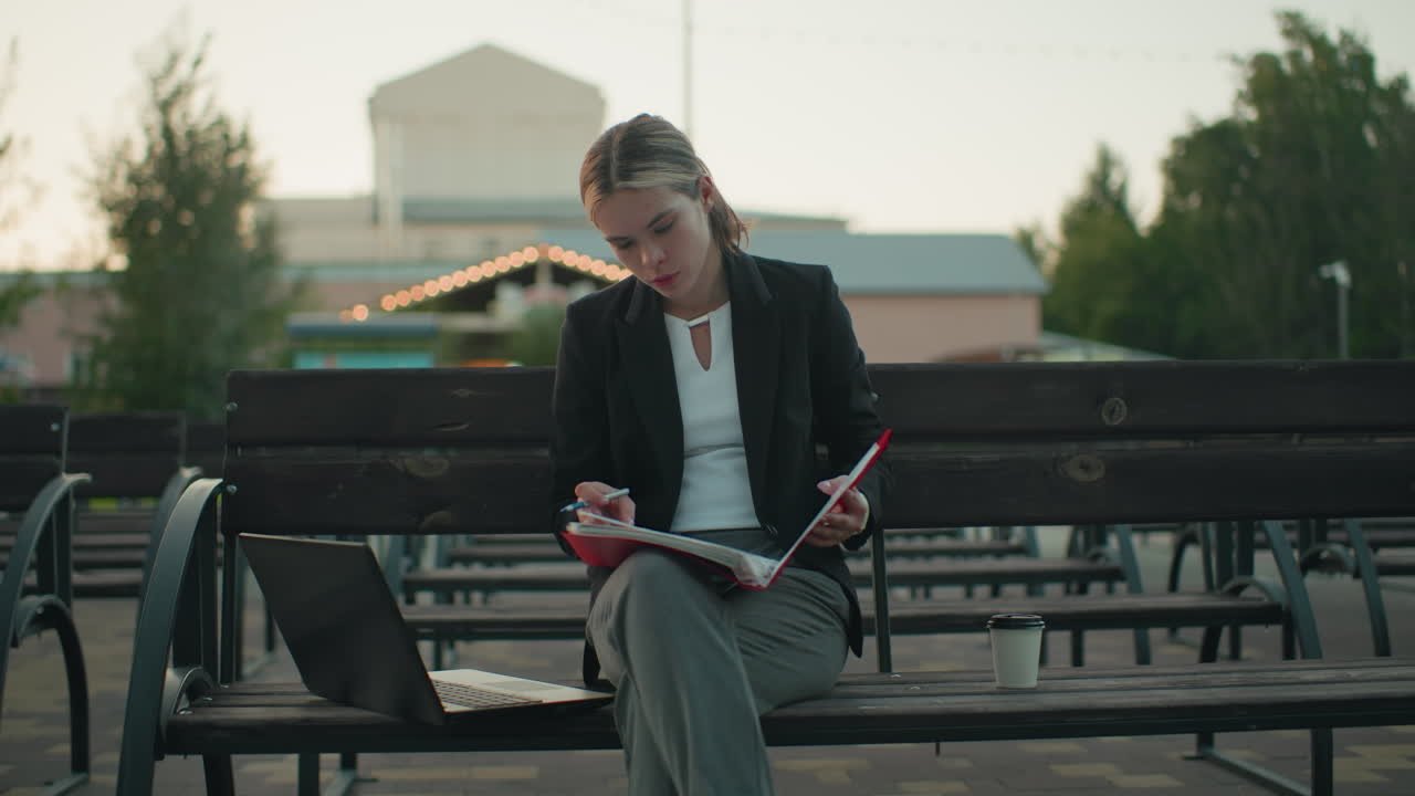 Young girl seated outdoor deeply focused on her book holding open folder with pen in hand, laptop on bench beside her and coffee cup nearby in urban setting with trees and building in background