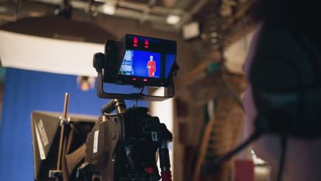 headset cameraman monitors camera during studio shoot, recording female presenter in red dress against blue screen, teleprompter visible, soft light shining, control gear filling production area