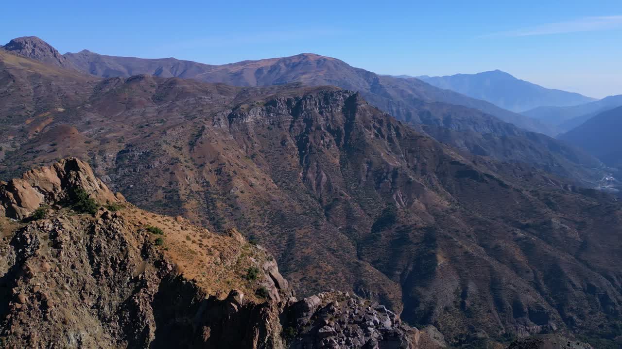 Aerial orbit around rocky peak with layered mountain ridges and valleys beyond
