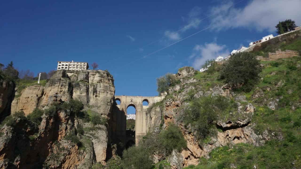 Time lapse showing stone bridge between rocky cliffs with fast moving clouds in blue sky and shadows shifting across landscape featuring hillside trees and scattered buildings