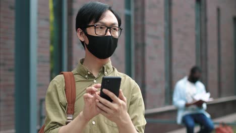 camera zooming on asian student wearing glasses and facial mask using his smartphone near the college