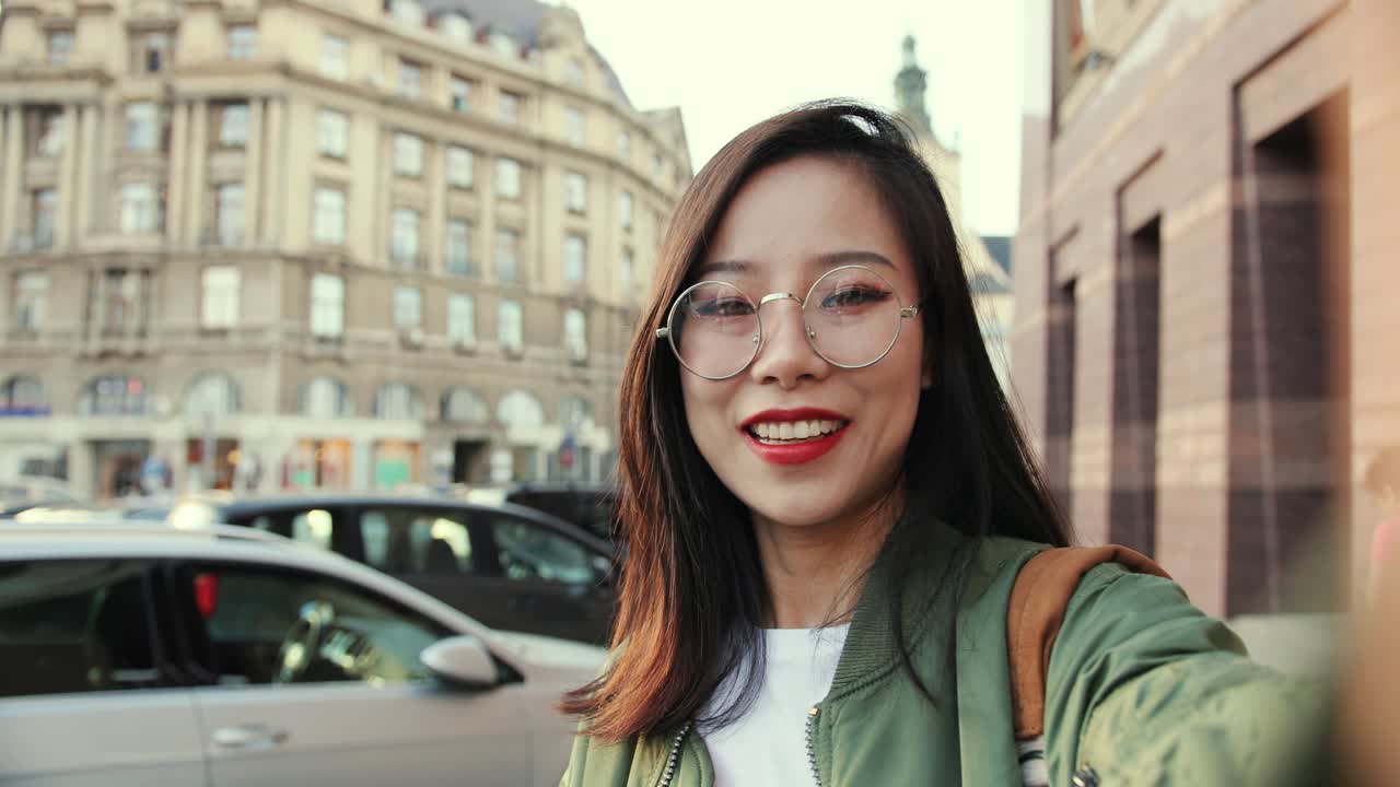 pov de la joven bastante alegre con gafas hablando con la cámara como tener videochat a través de la cámara web y mostrando el centro de la ciudad