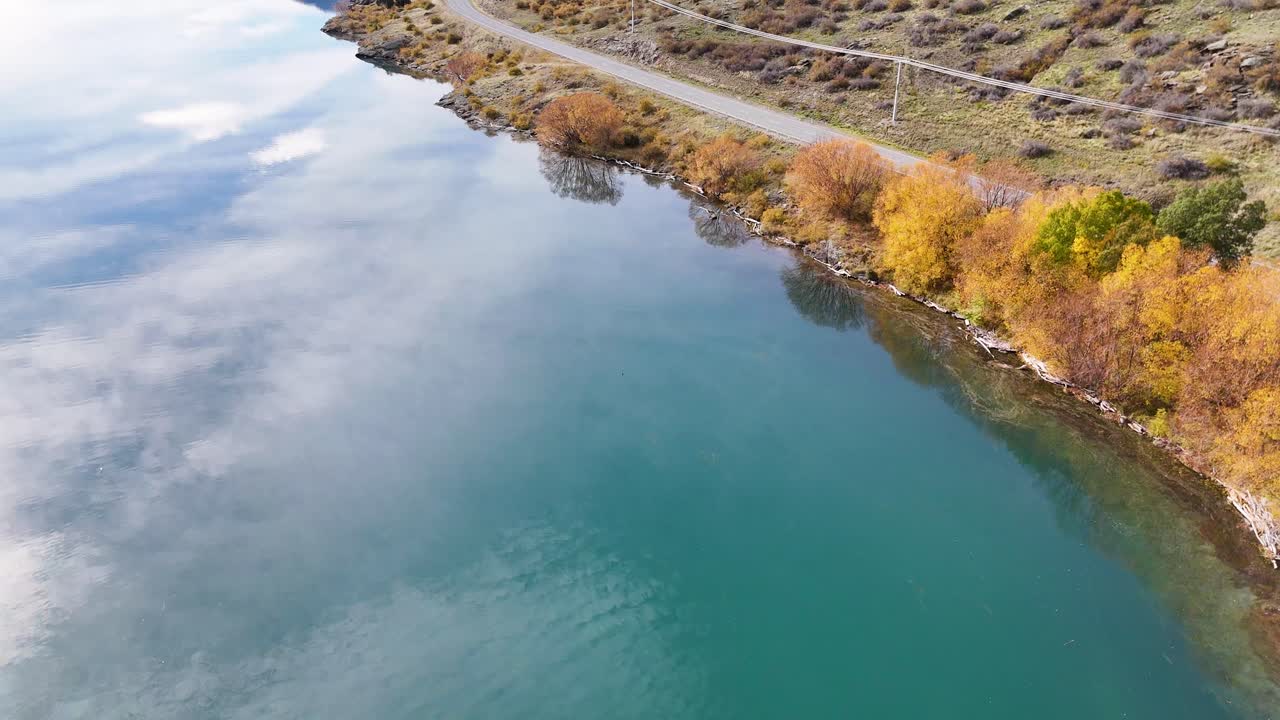 Drone captures serene autumn landscape with vibrant foliage along Lake Dunstan, Cromwell, New Zealand. Calm water reflects sky and trees