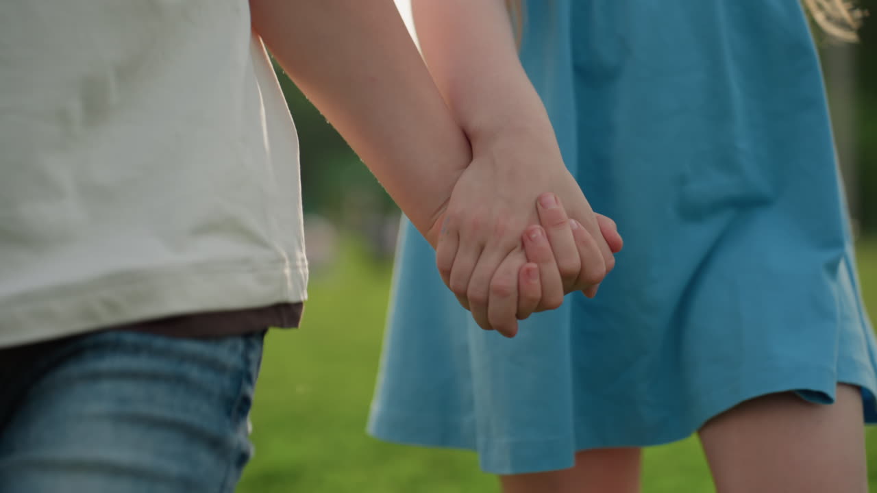 lower view of teenagers holding hands walking through playground grass, casual summer afternoon, friendship bond, relaxed sunlit park stroll, youthful connection, outdoor leisure, cheerful motion