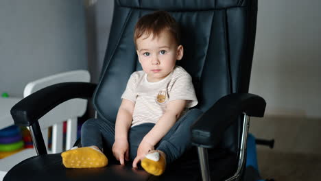 beautiful caucasian toddler boy sits on the big leather chair. kid looks at camera for a while with serious expression on his face and then smiles.
