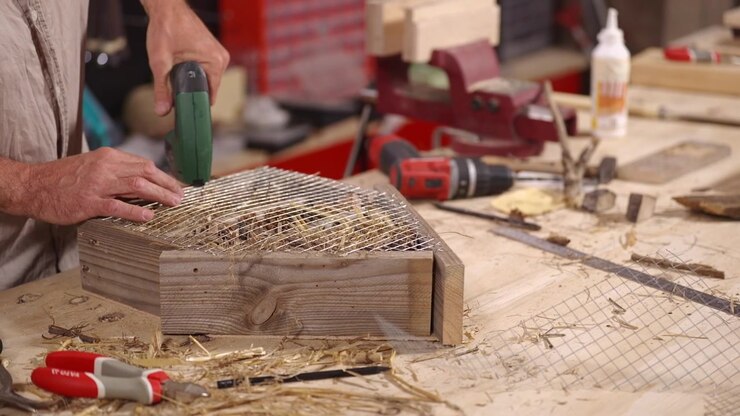 Hands secure wire mesh to a wooden insect hotel filled with natural materials using a staple gun at a workshop bench, creating shelter for garden wildlife.