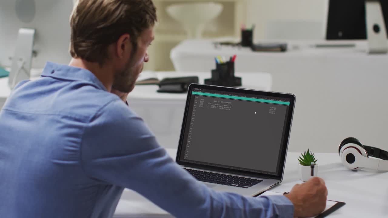 hombre caucásico sentado en un escritorio procesando en una computadora portátil y tomando notas