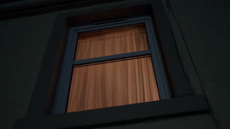 Looking-up-at-house-window-from-street-below,-at-night-with-warm-light-behind-curtain