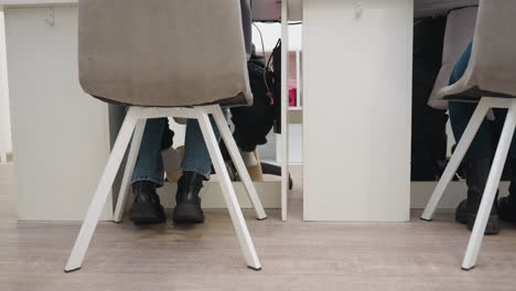 lower view showing two customers sitting at manicure desk inside nail salon, both wearing jeans and black boots, with legs visible beneath white furniture, capturing salon floor and modern setting