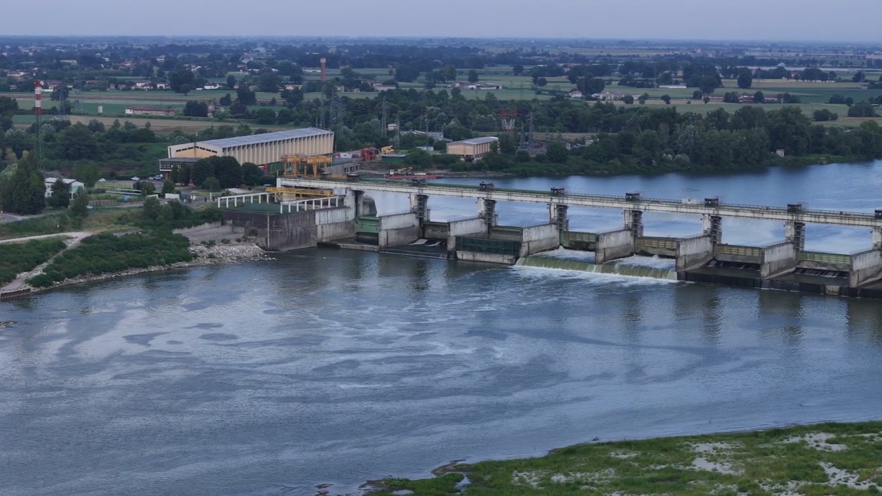 La diga di Isola Serafini regola il fiume Po vicino a Monticelli d'Ongina, Piacenza, con paratoie di sfioro, infrastrutture idroelettriche e terreni agricoli che circondano il complesso, in una ripresa panoramica.