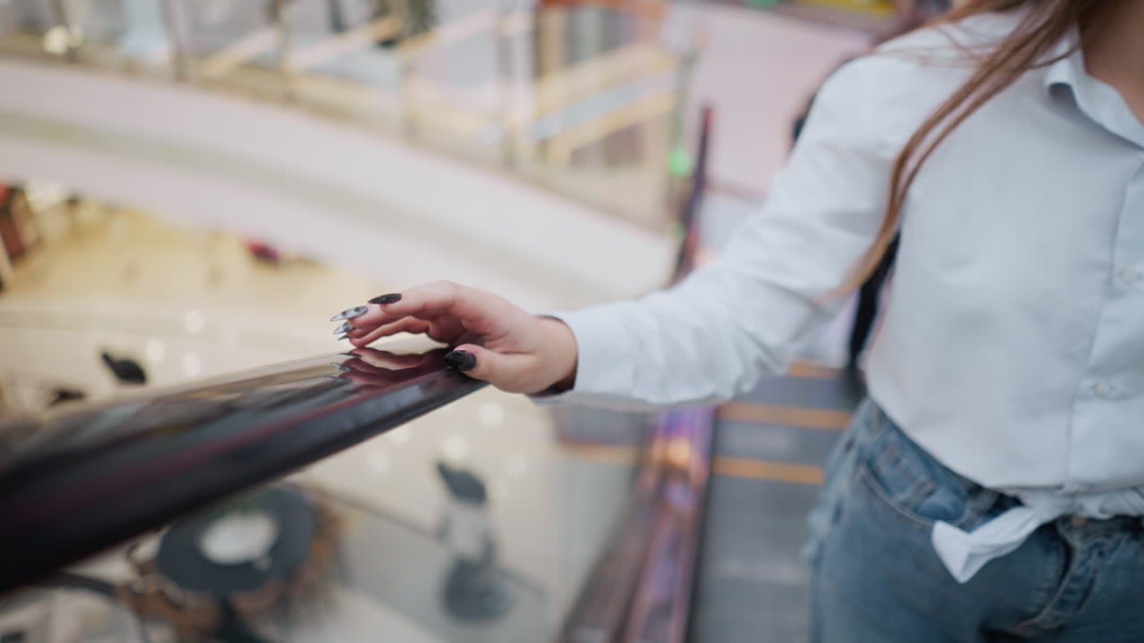 검은색 은 손을 가진 여성이 하강하는 동안 에스컬레이터 드레일을 부드럽게 니다. 그녀는 색 블라우스와 청바지를 입고 있습니다. 부드러운 초점의 배경은 흐릿한 쇼핑몰과 함께 현대적인 쇼핑몰을 포착합니다.