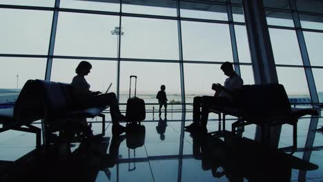 Una-Mujer-Con-Un-Niño-Esperando-Su-Vuelo-Se-Sientan-En-Sillas-En-Las-Siluetas-De-La-Terminal-Del-Aeropuerto