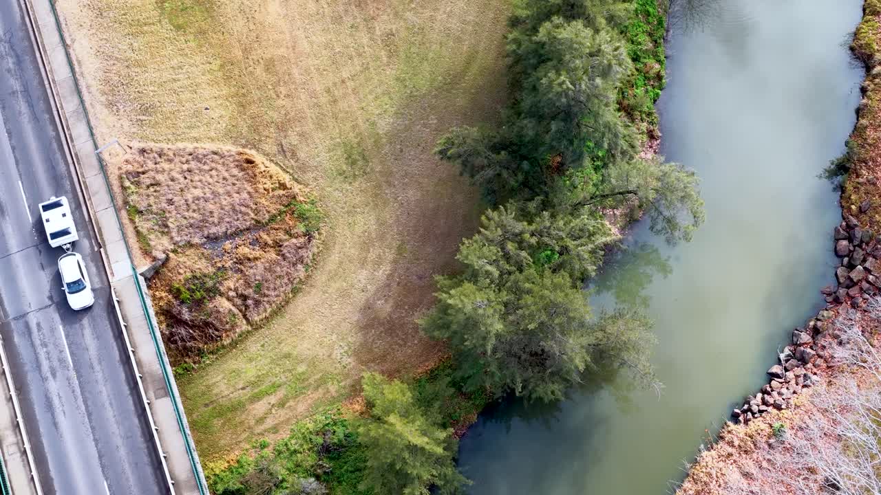 Drone footage tracks a white vehicle crossing a rural bridge above a winding river, surrounded by grassy banks and trees in daylight