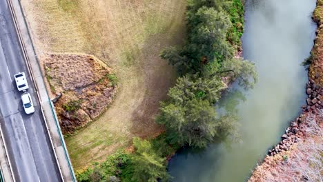 drone footage tracks a white vehicle crossing a rural bridge above a winding river, surrounded by grassy banks and trees in daylight