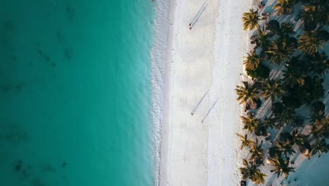 Perfekter-Flug-Aus-Der-Vogelperspektive-Drohnenaufnahme-Von-Menschen-Mit-Langen-Schatten-Auf-Weißem-Sand-Paradies-Traumstrand-Wellenküste-Zanzibar,-Afrika-Tansania-2019-Cinematic-1080,-60p-Von-Philipp-Marnitz