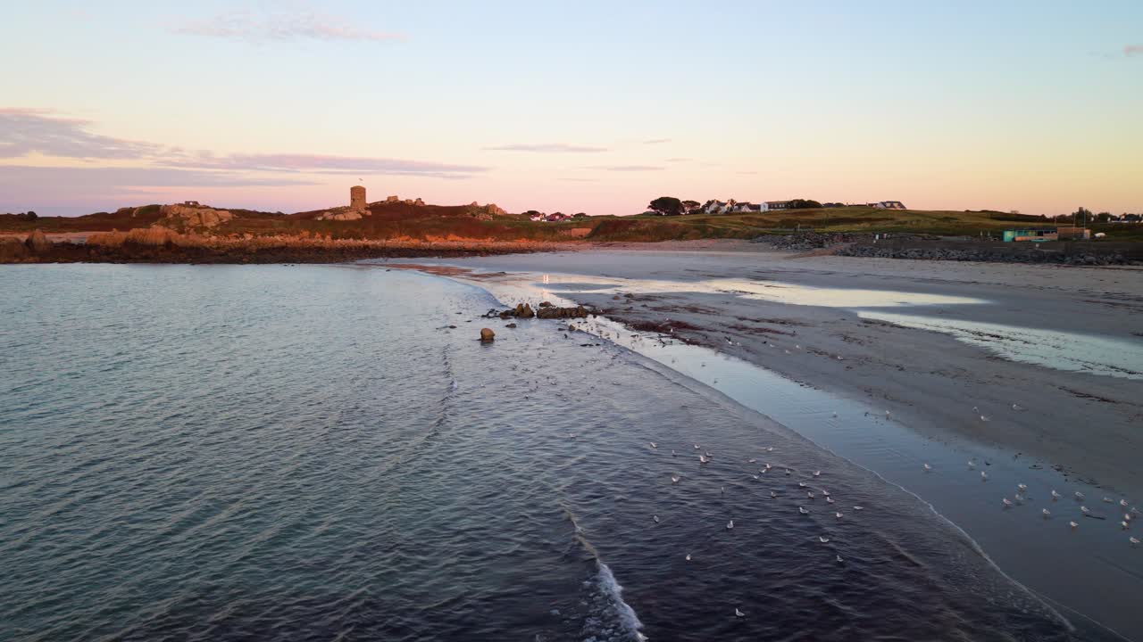 Lancresse Bay Guernsey, långsam låg flygning längs strandlinjen av sandstrand med klippformationer under den gyllene timmen med lugna, kluckande vågor, med Martellotornet gyllene på den bortre stranden och fåglar på strandlinjen.