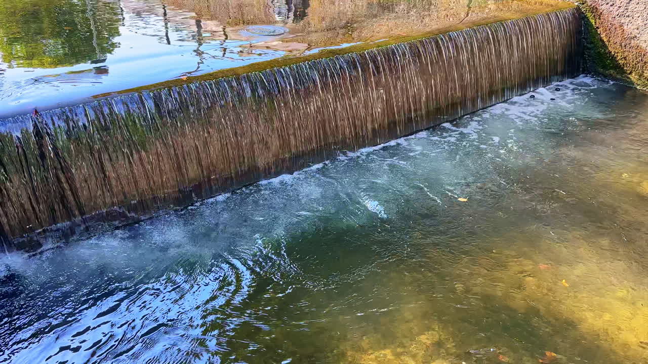 L'acqua scorre dolcemente su una sporgenza di pietra creando increspature e riflessi in un sereno ambiente naturale vicino a un parco durante una giornata di sole.