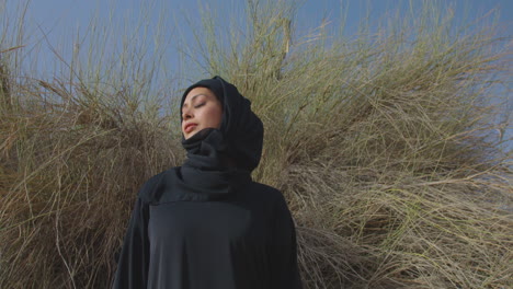 retrato de una hermosa mujer musulmana con vestido tradicional e hiyab parada cerca de un arbusto del desierto y sonriendo a la cámara