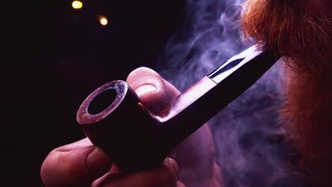 closeup: man with bushy beard puffs smoke from pipe in backlit room