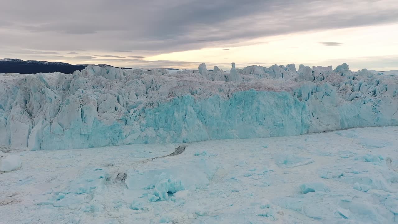 Drone shot of a massive blue glacier wall in Greenland, revealing dramatic textures, deep ice layers, and the vast power of the Arctic wilderness under a cloudy sky