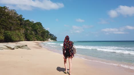 a tall blond girl walks along a deserted beach in the andaman islands in india with forest lining the sand and blue sky