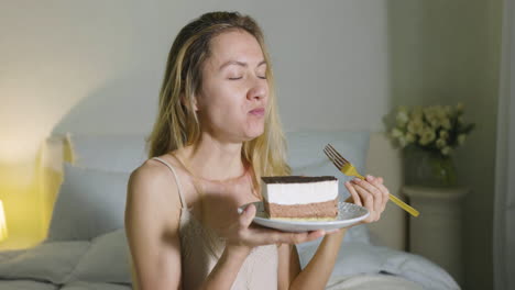 chica rubia sosteniendo y comiendo un trozo de pastel sentado en la cama 1