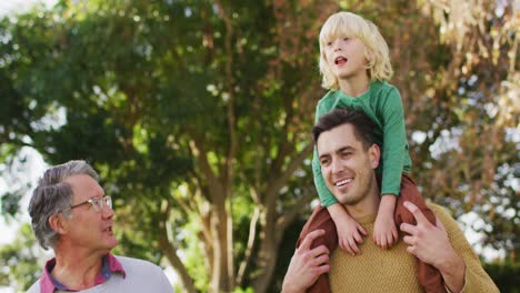video de un padre caucásico sonriente llevando a su hijo sobre los hombros caminando con su abuelo en el jardín