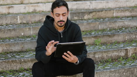 Young-Arabic-handsome-man-with-dark-curly-hair-and-beard-in-black-hoodie-sitting-on-stairs-outside