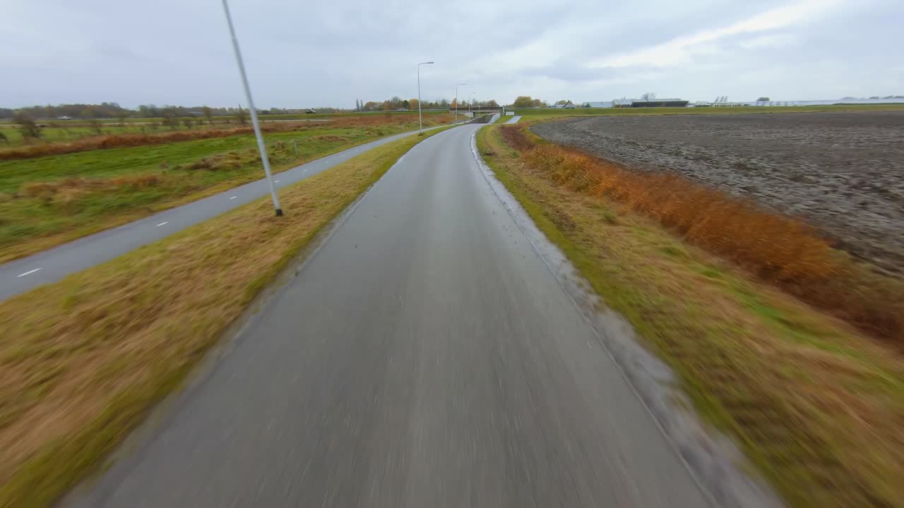 Drone following a small road towards a busy elevated main road, surrounded by an agricultural area