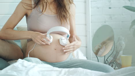 unrecognized pregnant woman holding headphones on her belly at home.