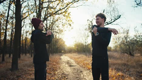 a man with curly hair and a beard in a black sports uniform together with his friend in a red cap are doing sports and warming up before jogging in an autumn park in the morning