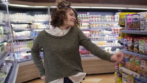 retrato de una mujer joven con camisa y cabello rizado bailando de pie en el pasillo de la tienda de comestibles. mujer emocionada divirtiéndose, bailando en el supermercado, sonriendo. cámara lenta