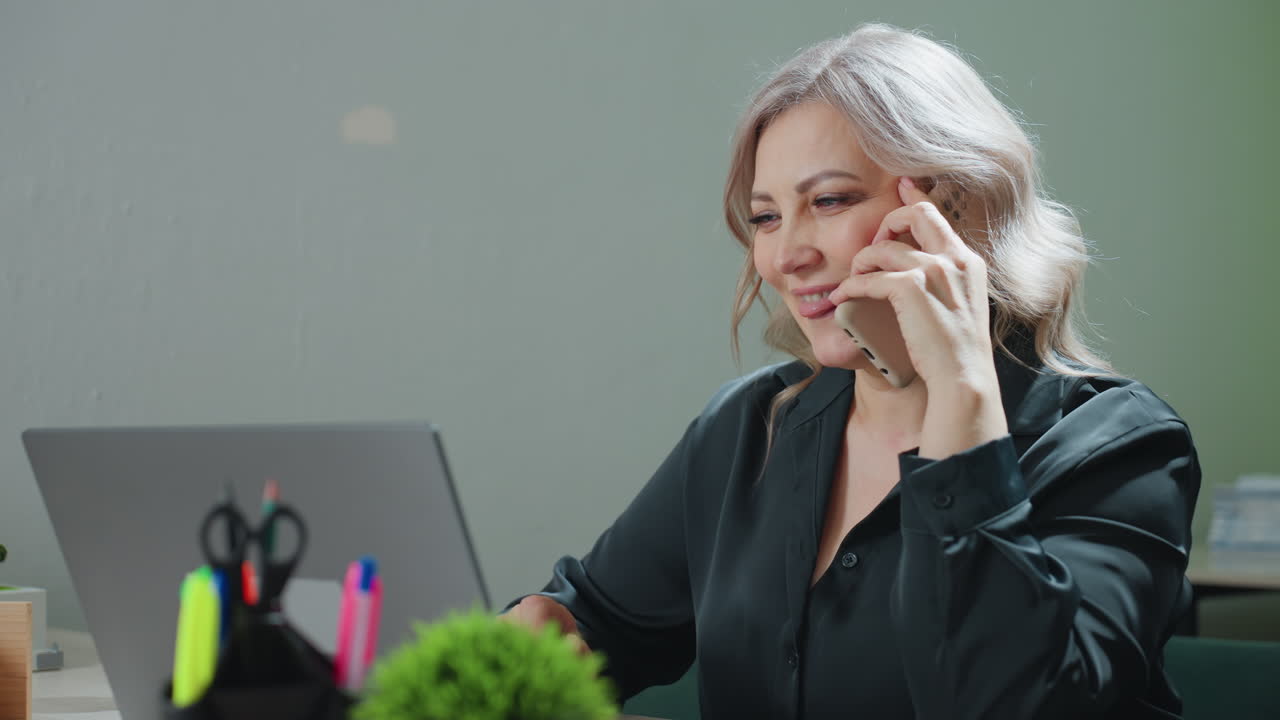 Confident professional lady with warm demeanor engaged in client call while working on laptop at tidy office desk, focused and multitasking with thoughtful expression in soft daylight environment