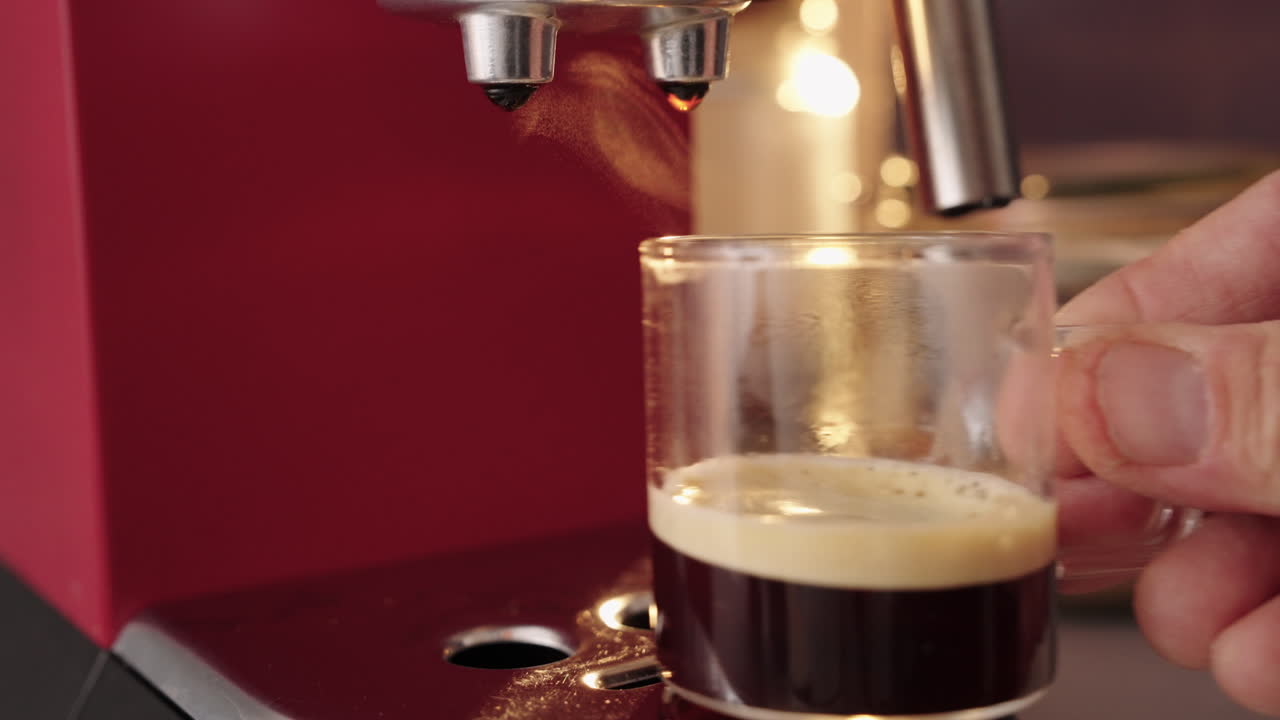Primer plano de un espresso recién hecho en una taza de cristal transparente bajo una cafetera roja en una cocina casera. El vapor se eleva con luz natural mientras una mano retira la taza. Un café matutino real y cotidiano