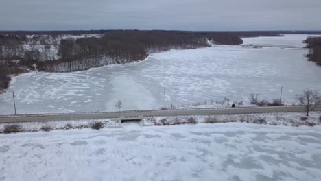 4k-Aerial-footage-of-a-small-icy-frozen-lake-in-Michigan,-USA-taken-on-a-cold-winter-day-outdoors-moving-away-from-a-small-bridge-in-a-rural-area