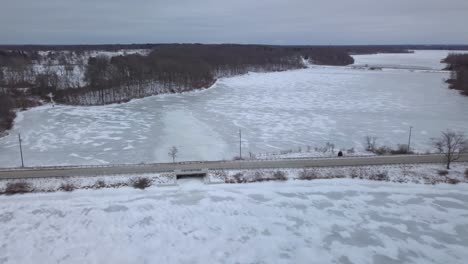 4k aerial footage of a small icy frozen lake in michigan, usa taken on a cold winter day outdoors moving away from a small bridge in a rural area