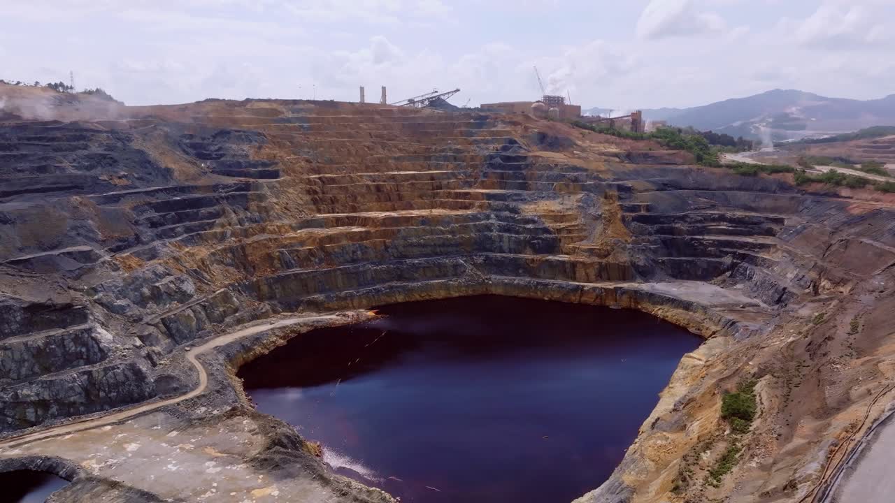 Dark and reflective water of open-pit mine lake, terraced walls of excavation site, Barrick Gold mining Cotuí, Dominican Republic. Aerial forward