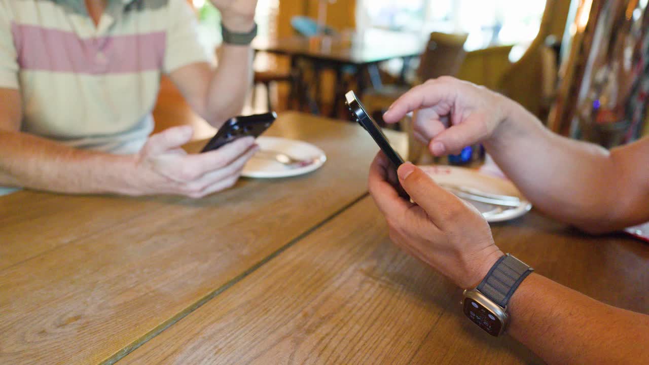 Two people interact with smartphones at a wooden table in a bright, casual restaurant setting